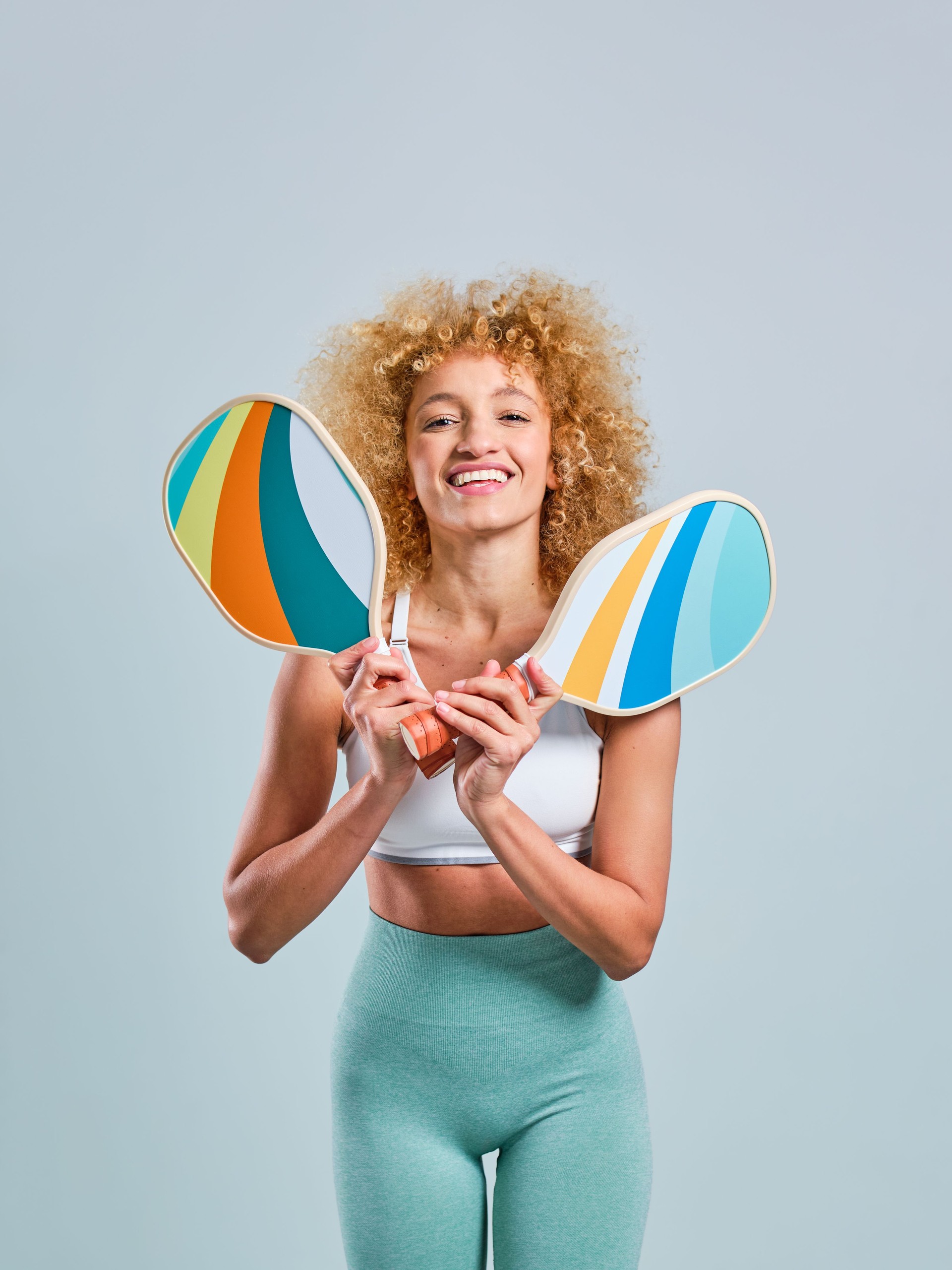 Young Brazilian girl with blonde afro hair facing front smiling with two crossed pickleball paddles